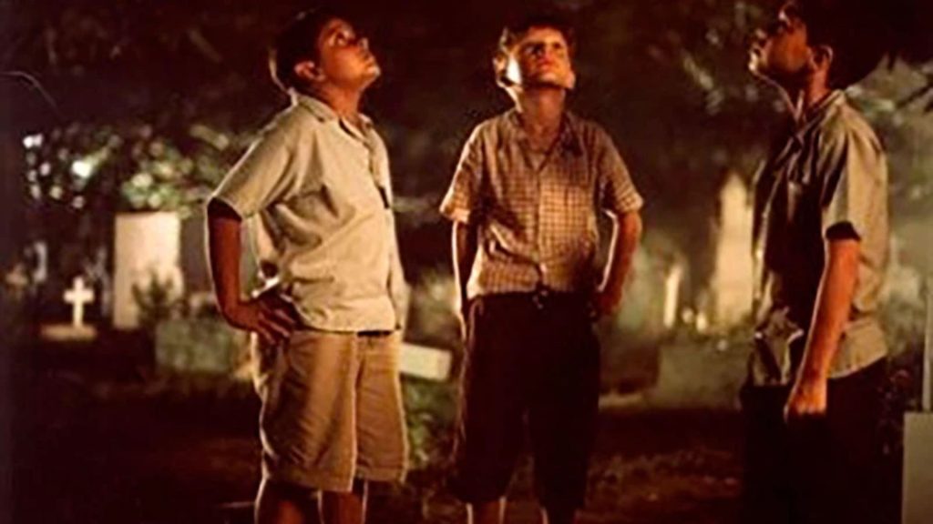 Tres niños observan el cielo en un cementerio, rodeados de tumbas, en una escena nocturna.
