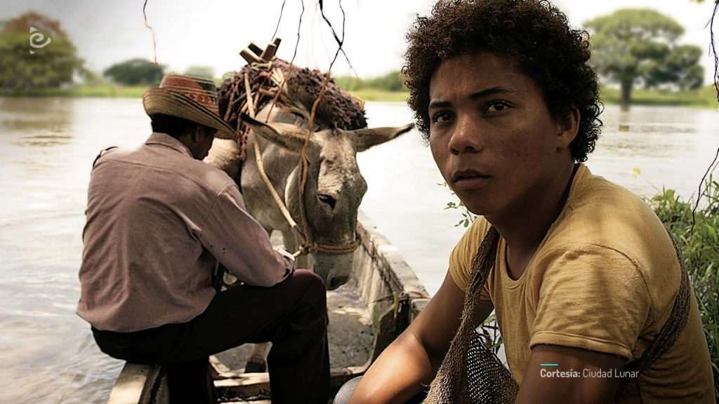 Joven con camiseta amarilla a orillas del río, junto a un burro y un hombre mayor en una canoa.