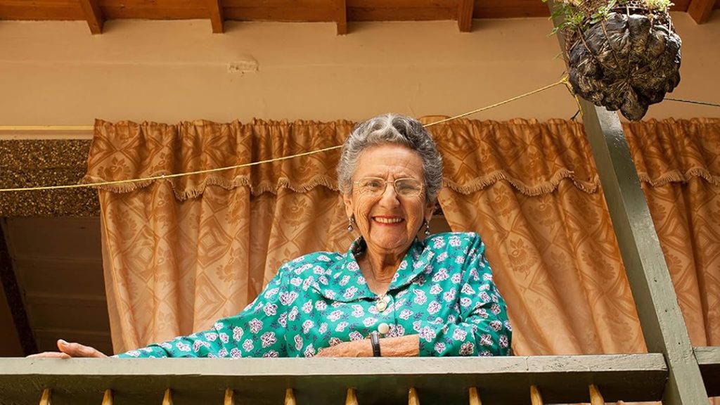 Mujer en Jericó mayor sonriente asomada desde un balcón, vestida con camisa verde estampada.