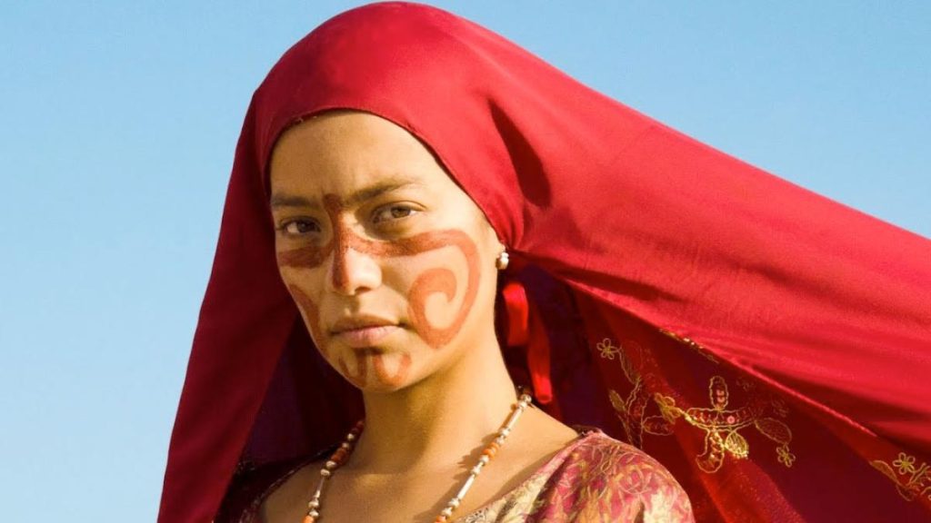 Mujer wayuu con pintura facial roja y velo rojo ondeando al viento, bajo el cielo azul-Pájaros de verano