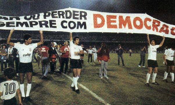 Jugadores del Corinthians sostienen una larga pancarta con el lema “Ganhar ou perder, mas sempre com Democracia” en el campo, rodeados de prensa y aficionados.