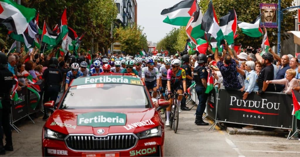 Aficionados con banderas palestinas a ambos lados de la carretera durante la Vuelta a España 2025; paso del pelotón y coche director. Un rechazo al sportswashing en el ciclismo.