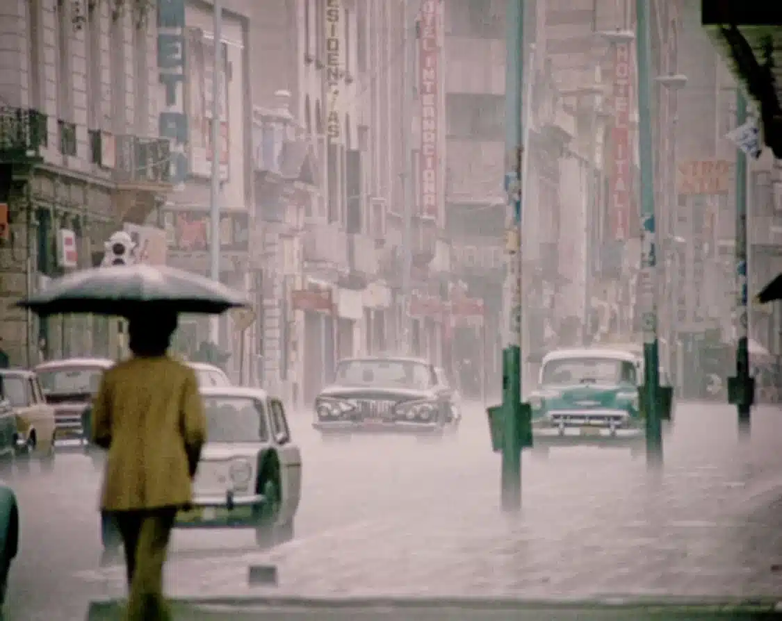 Fotografía histórica de la carrera Séptima de Bogotá bajo la lluvia, con una persona bajo un paraguas y carros de época