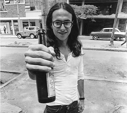 Andrés Caicedo joven, con gafas y el pelo largo, sonríe y extiende hacia la cámara una botella de cerveza Pola